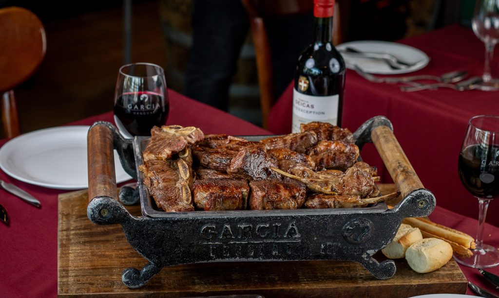 Brasero de carne García con cortes a la parrilla, vino tinto y pan en mesa servida.
